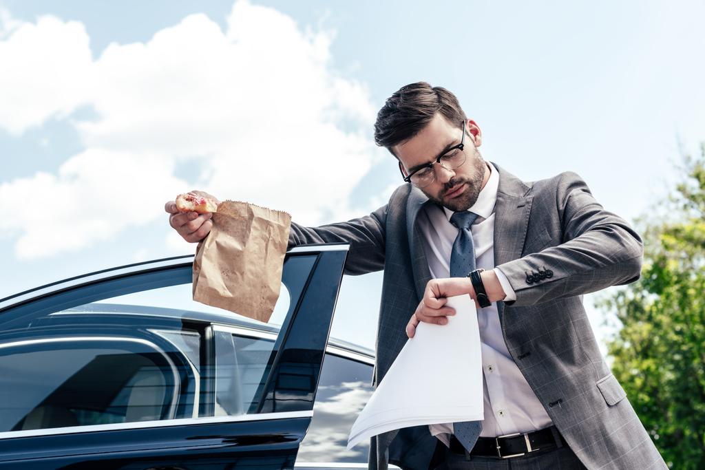 бизнесмен с отнять еду и документы в руках проверки времени, стоя у автомобиля на улице
 - Фото, изображение