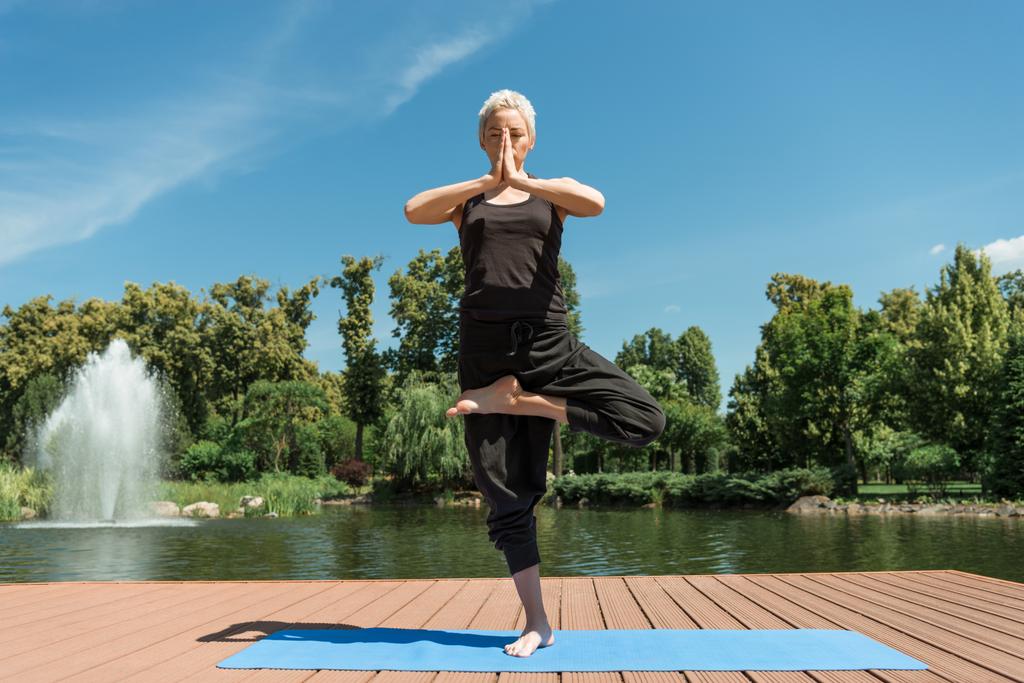 woman practicing yoga in Tree pose (Vrksasana) on yoga mat near river in park - Photo, Image