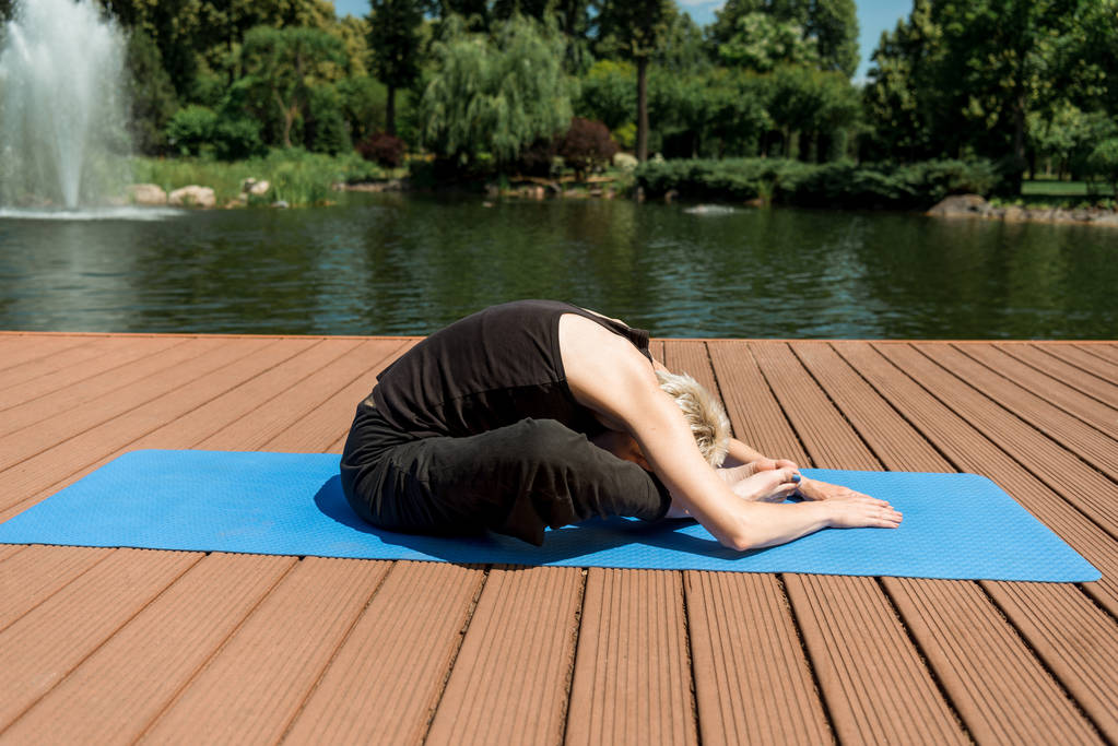 vista laterale della donna che pratica yoga in posa Wide Child (Balasana) sul tappeto yoga vicino al fiume nel parco
 - Foto, immagini