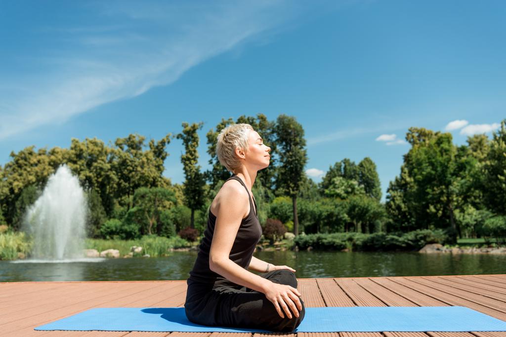 Zijaanzicht van gelukkige vrouw praktizerende yoga in lotus vormen op yoga mat in de buurt van rivier in park - Foto, afbeelding
