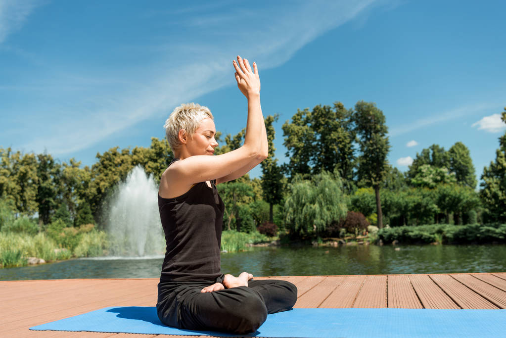 vista lateral da mulher praticando ioga em pose de lótus e mãos em gesto namaste no tapete de ioga perto do rio no parque
 - Foto, Imagem