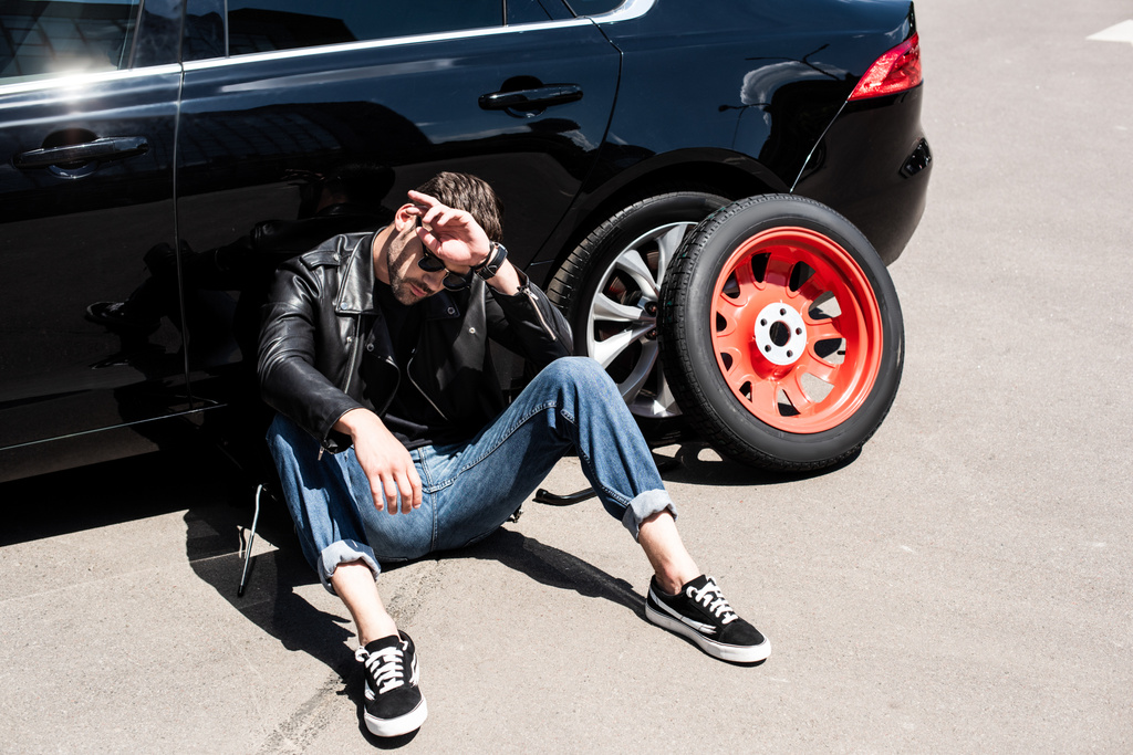 stylish tired man in sunglasses wiping forehead near tools, tire and broken car at street - Photo, Image