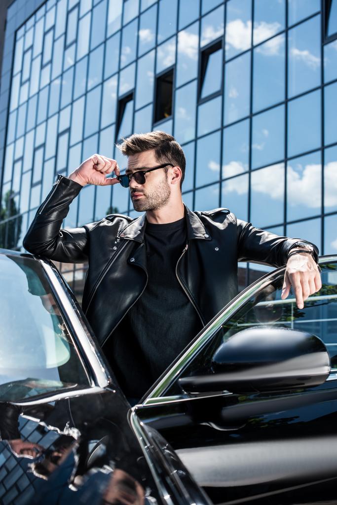 стильний чоловік у шкіряній куртці та сонцезахисних окулярах, що дивиться далеко біля машини на вулиці
 - Фото, зображення