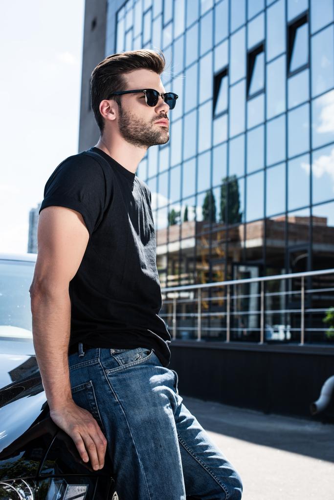 Вид сбоку стильного молодого человека в солнцезащитных очках, стоящего возле своей машины на улице
 - Фото, изображение
