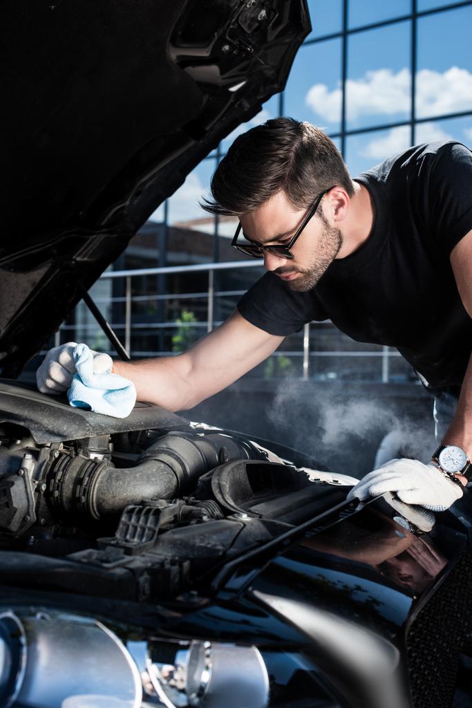 handsome young man in working gloves holding rag and looking at engine of broken car with smoke coming out  - Photo, Image