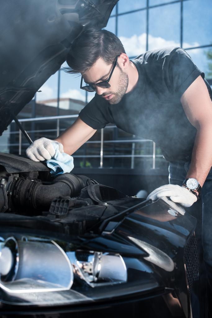 jonge man in werkhandschoenen probeert te herstellen van de motor van gebroken auto met rook uit  - Foto, afbeelding