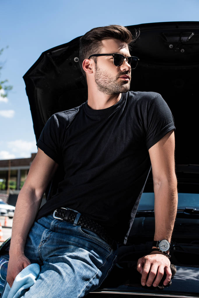 Вид сбоку молодого человека в солнцезащитных очках с тряпкой возле машины с открытым капотом на улице
 - Фото, изображение