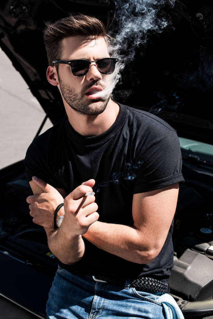 Stilvoller junger Mann mit Sonnenbrille raucht Zigarette, während er neben kaputtem Auto mit geöffneter Motorhaube steht  - Foto, Bild