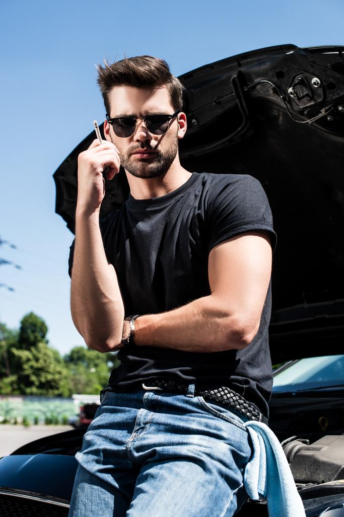 κομψό νεαρός με γυαλιά ηλίου κάπνισμα τσιγάρου κοντά σπασμένο αυτοκίνητο με ανοιγμένο καπό  - Φωτογραφία, εικόνα