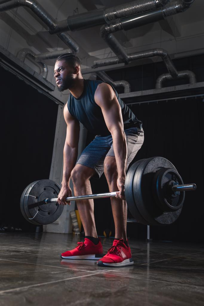 low angle view of muscular african american sportsman lifting barbell and looking away in gym  - Photo, Image