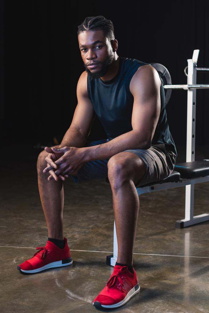 Ganzkörperansicht des jungen afrikanisch-amerikanischen Sportlers, der im Fitnessstudio sitzt und in die Kamera schaut - Foto, Bild