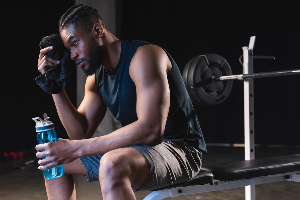 deportista afroamericano cansado sosteniendo la toalla y la botella de agua mientras está sentado en el gimnasio
 - Foto, imagen