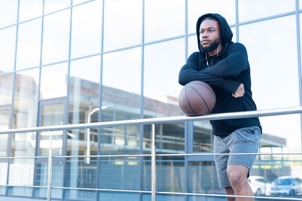 συλλογισμένος αφρικανική αμερικανική άνθρωπος στα ακουστικά ακουμπά στα κάγκελα με μπάλα μπάσκετ και αναζητούν μακριά - Φωτογραφία, εικόνα