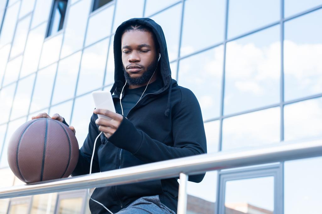niski kąt widzenia sportive african american man w słuchawki za pomocą smartfonu, stojąc z piłkę do koszykówki na ulicy - Zdjęcie, obraz