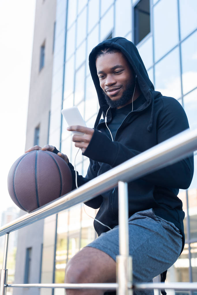 усміхнений афроамериканський чоловік в навушниках, використовуючи смартфон, тримаючи баскетбол на вулиці
 - Фото, зображення