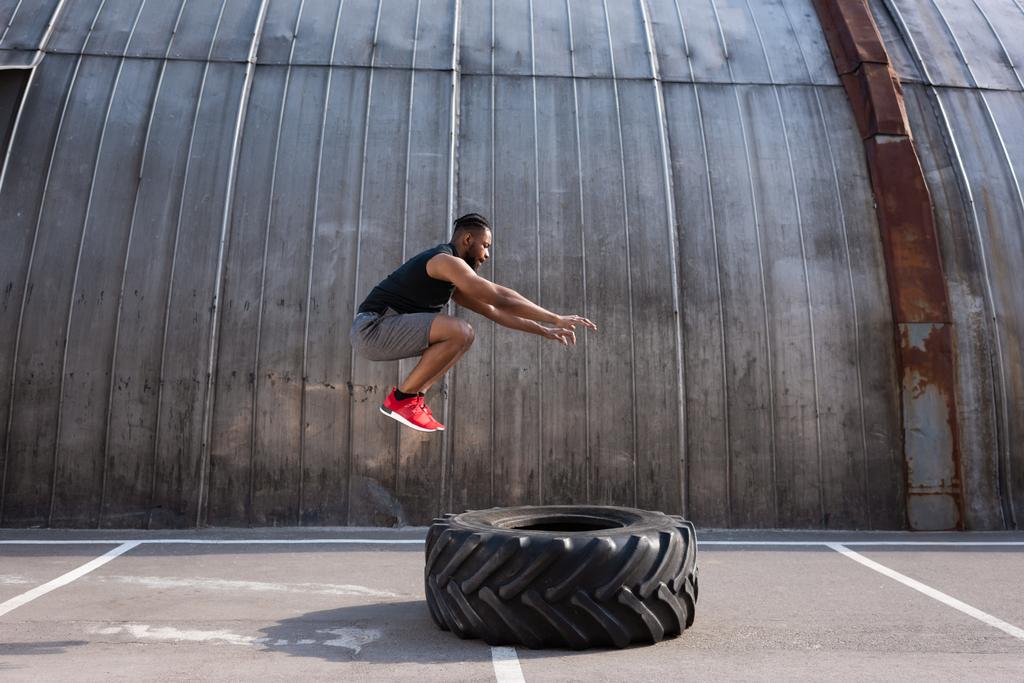muscular african american sportsman jumping while training with tyre on street  - Photo, Image