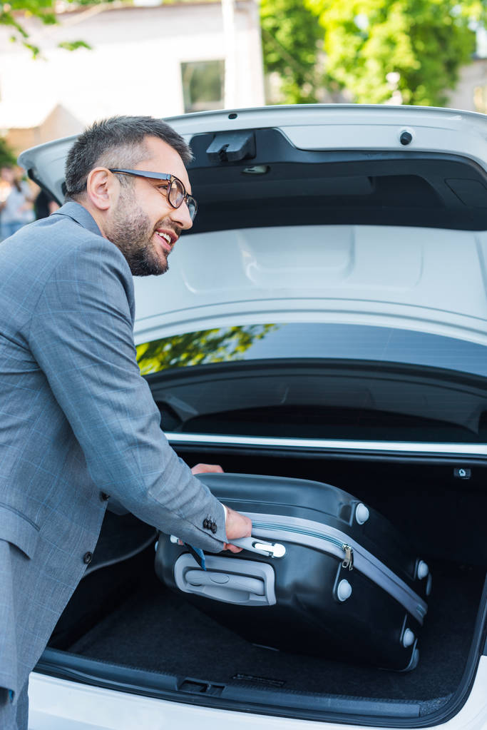 side view of smiling businessman putting luggage into car on parking - Photo, Image