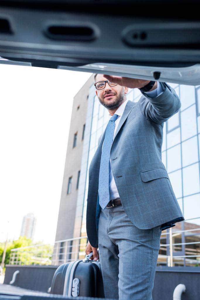 επιχειρηματίας σε γυαλιά με αποσκευές στέκεται στο αυτοκίνητο στο χώρο στάθμευσης - Φωτογραφία, εικόνα