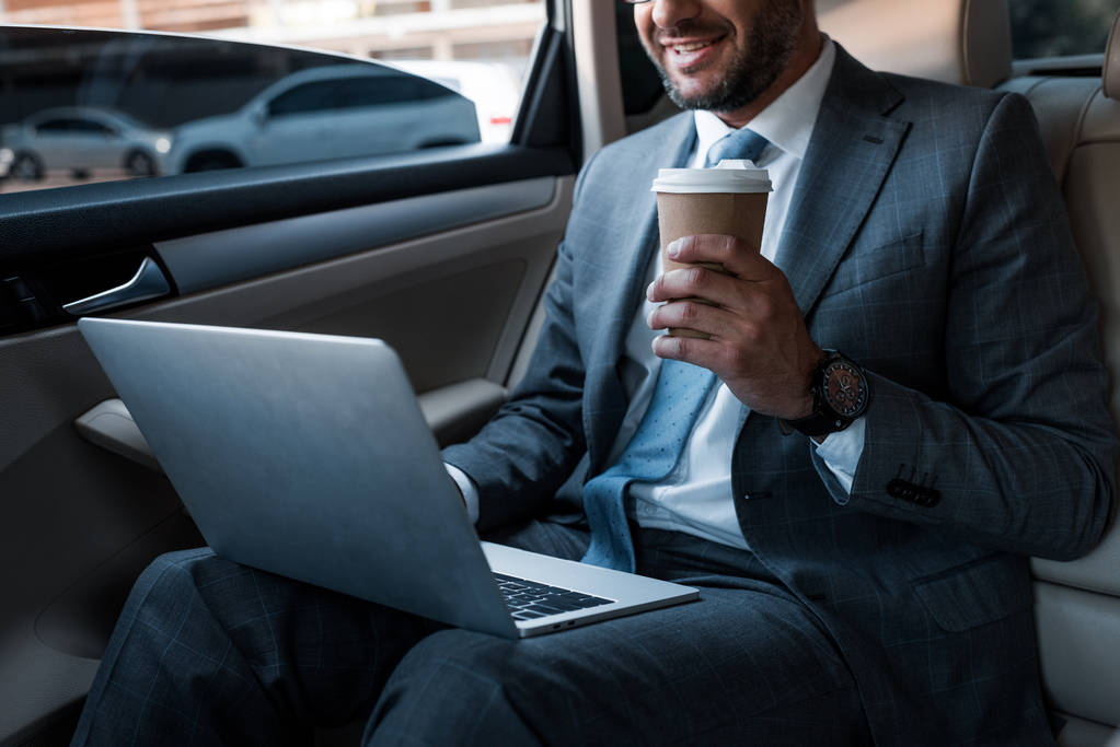 plan recadré d'homme d'affaires souriant avec du café pour aller à l'aide d'un ordinateur portable sur le siège arrière dans la voiture
 - Photo, image
