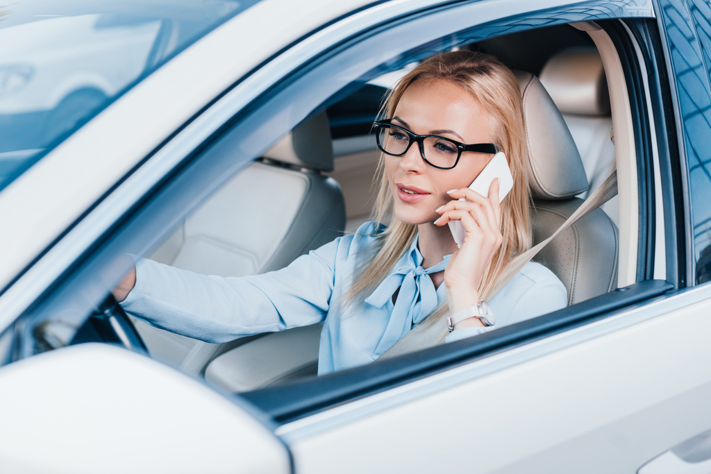 Блондинка-бизнесвумен в очках разговаривает по смартфону за рулем автомобиля
 - Фото, изображение
