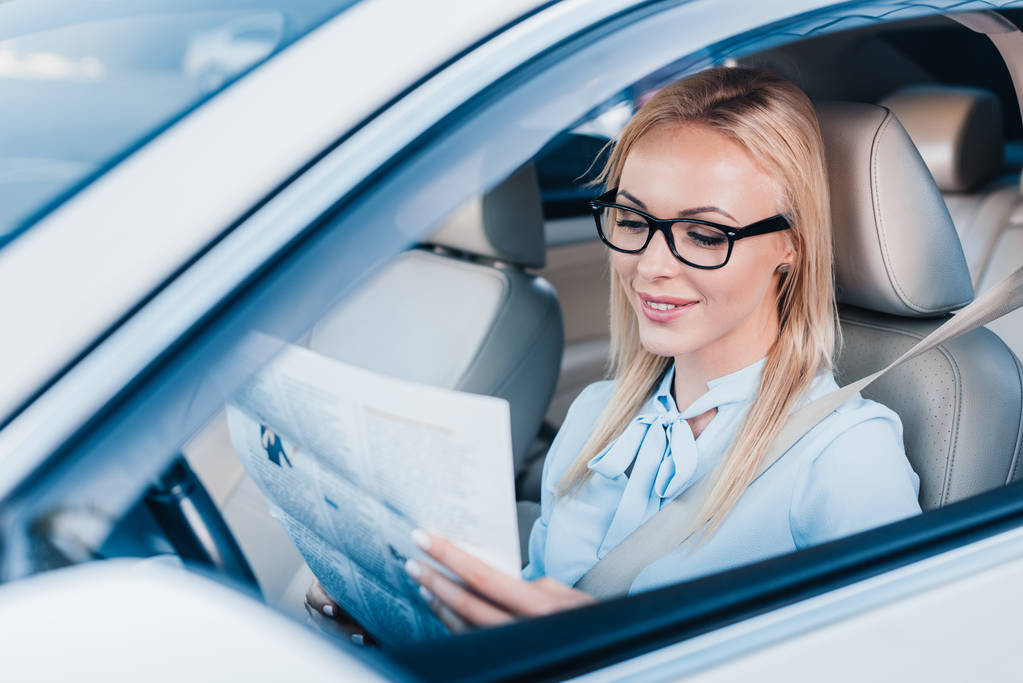 hymyilevä liikenainen lukee sanomalehteä autossa
 - Valokuva, kuva