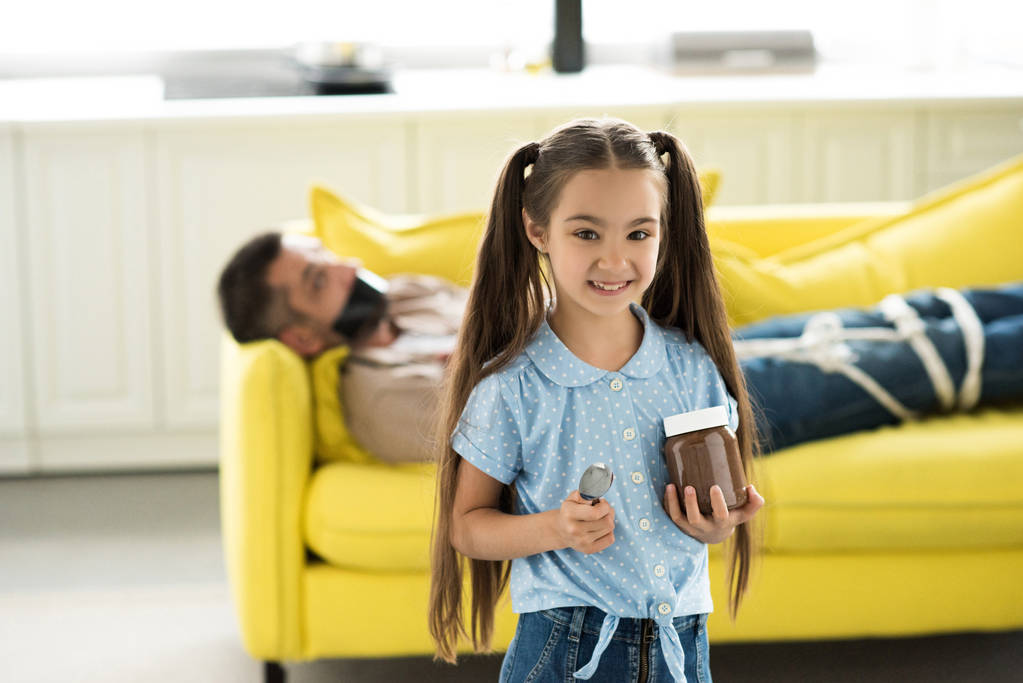 ο πατέρας που βρίσκεται δεμένο στον καναπέ και χαμογελαστά κόρη κρατώντας σοκολάτας στο σπίτι - Φωτογραφία, εικόνα