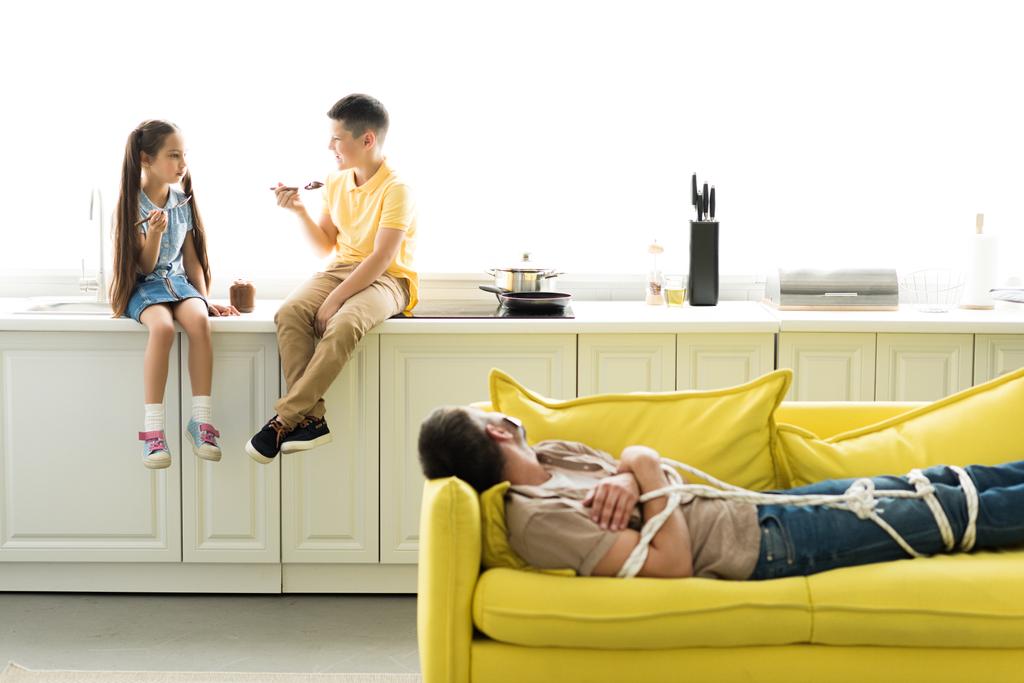 crianças comendo chocolate na cozinha e pai deitado amarrado no sofá
 - Foto, Imagem