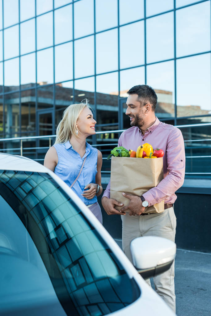 portrait of married couple with paper bag full of healthy food on parking near car - Photo, Image