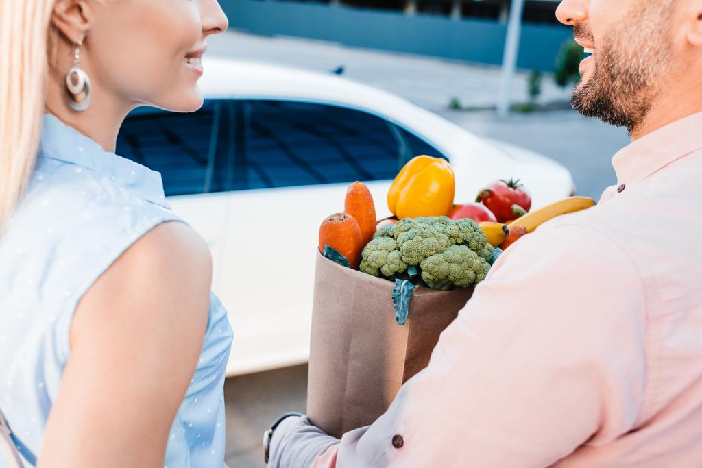 περικοπεί shot από παντρεμένο ζευγάρι με χάρτινη σακούλα γεμάτη από υγιεινά τρόφιμα στο χώρο στάθμευσης κοντά στο αυτοκίνητο - Φωτογραφία, εικόνα