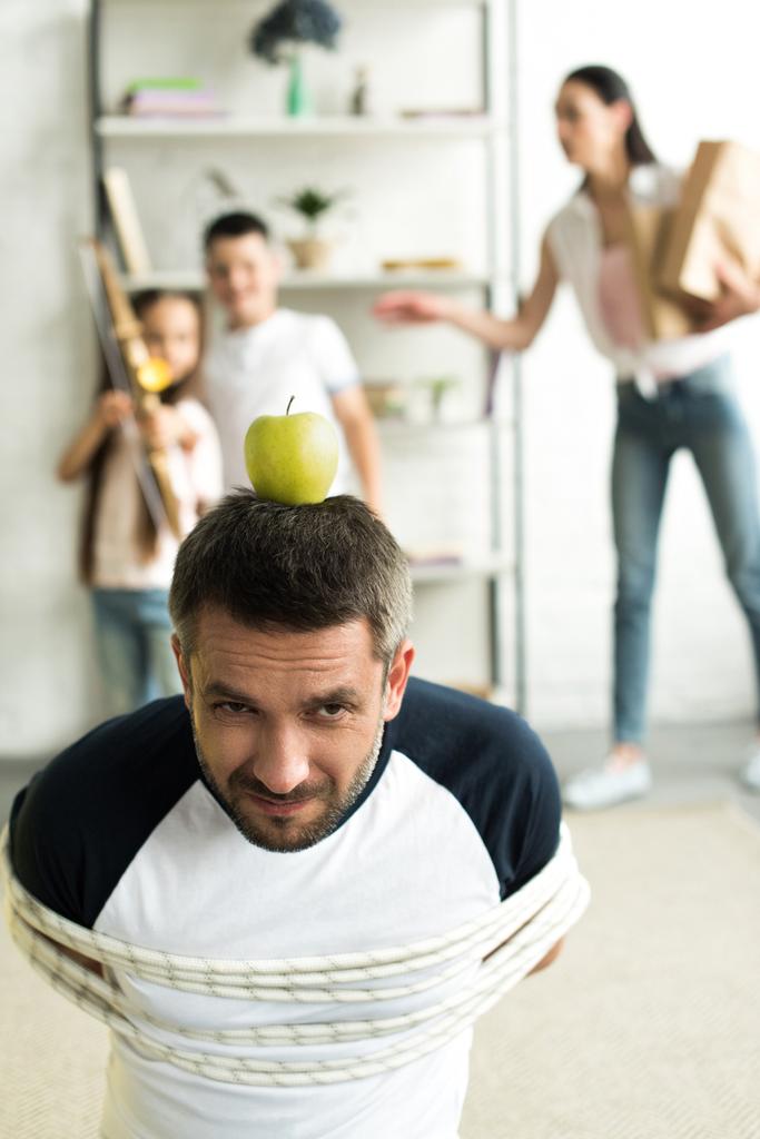 tied father sitting on floor with apple on head for target at home, parenthood concept - Photo, Image