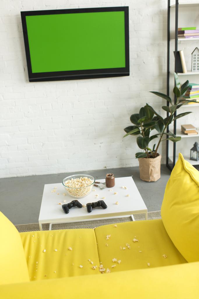 vista de ángulo alto del tazón de vidrio con palomitas de maíz y joysticks en la mesa cerca del sofá amarillo en la sala de estar
 - Foto, Imagen