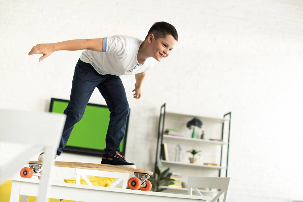 lachende vooraf adolescent jongen staande op skateboard op tafel thuis - Foto, afbeelding