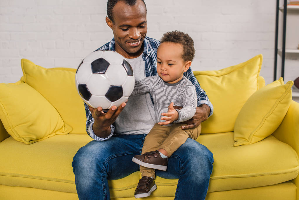 happy young father holding soccer ball while sitting with adorable little son on sofa - Photo, Image