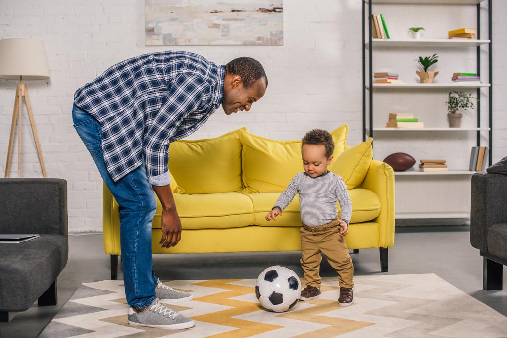 χαρούμενος νεαρός πατέρας βλέπει χαμογελαστός ο μικρός γιος παίζει με μπάλα ποδοσφαίρου στο σπίτι - Φωτογραφία, εικόνα