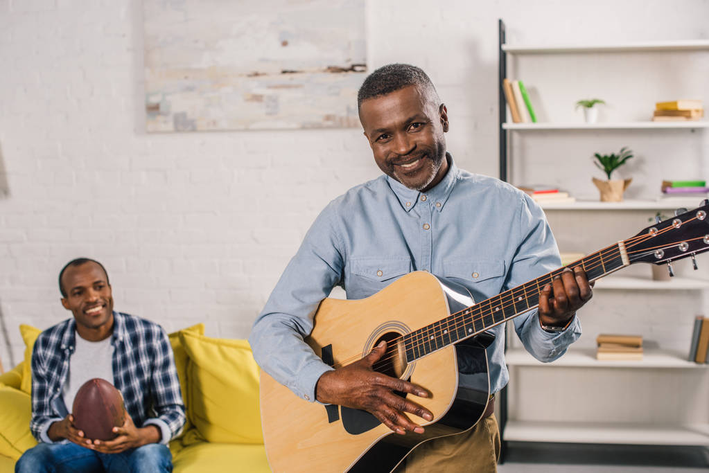 idősebb ember, akusztikus gitáron játszik, és a kamera, míg a felnőtt fia gazdaság rögbi labda mögött mosolygó - Fotó, kép