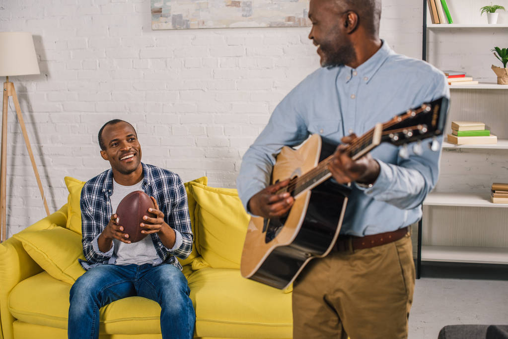 oříznutý snímek starší muž hraje na akustickou kytaru a při pohledu na usmívající se dospělého syna doma drží rugby míč - Fotografie, Obrázek