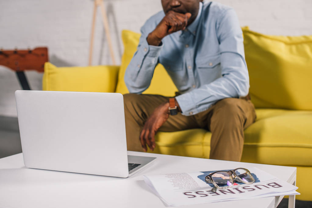 a táblázat és a vezető afrikai amerikai férfi, ülő-on dívány mögött laptop, szemüveg és újság kiadványról - Fotó, kép