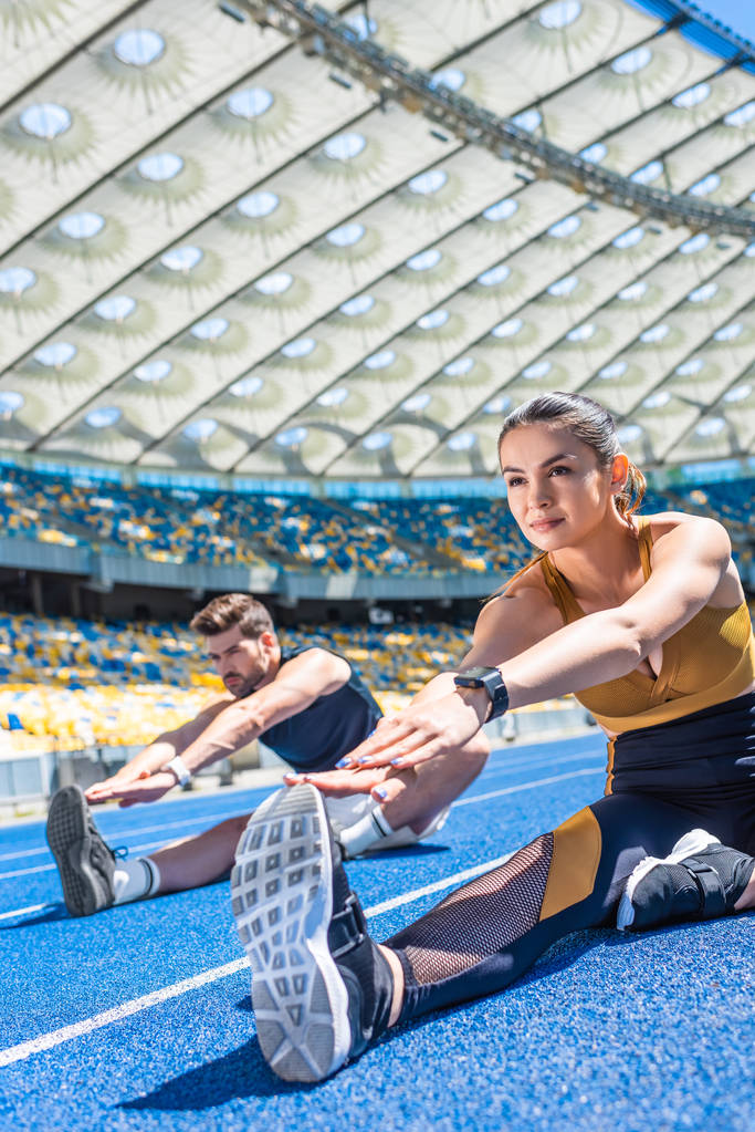 joven pareja atractiva sentado en pista de atletismo y estiramiento en el estadio deportivo
 - Foto, Imagen