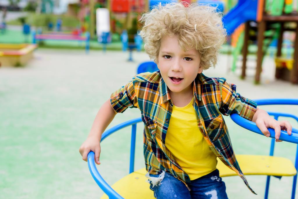 selective focus of curly little boy riding on carousel at playground  - Photo, Image