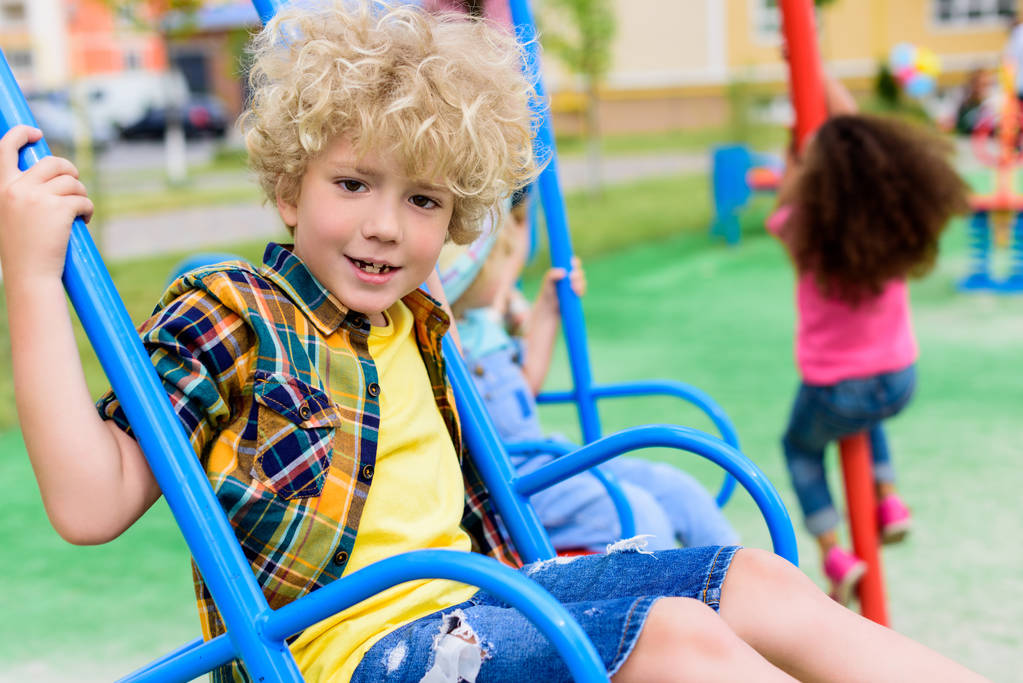 enfoque selectivo de niño rizado feliz montar en columpio en el patio de recreo
  - Foto, Imagen