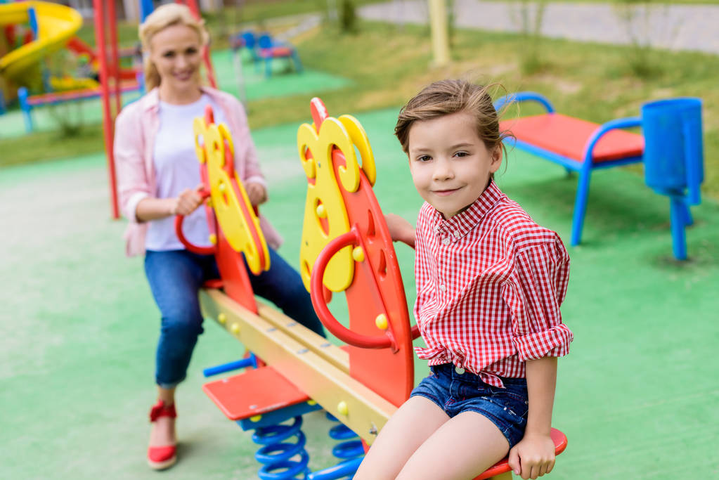 foco seletivo de uma criança adorável sentada em um cavalo de baloiço enquanto sua mãe está sentada atrás do playground
  - Foto, Imagem
