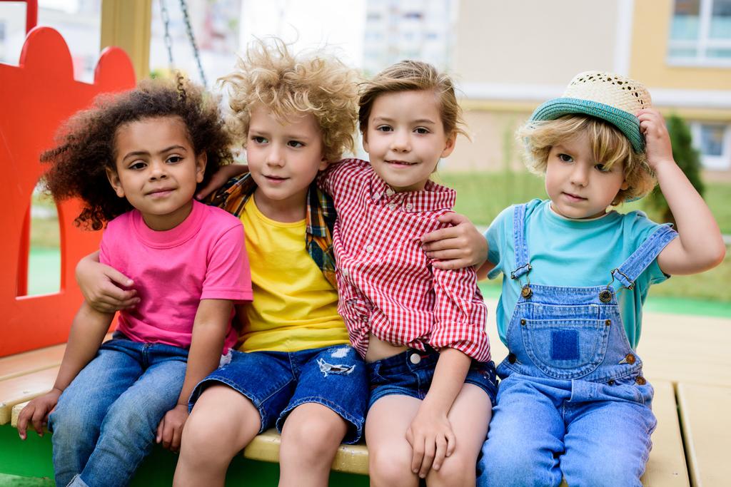 chłopiec kręcone, obejmującego dwa wielokulturowym dzieci podczas innych chłopiec siedzi w pobliżu plac zabaw dla dzieci  - Zdjęcie, obraz