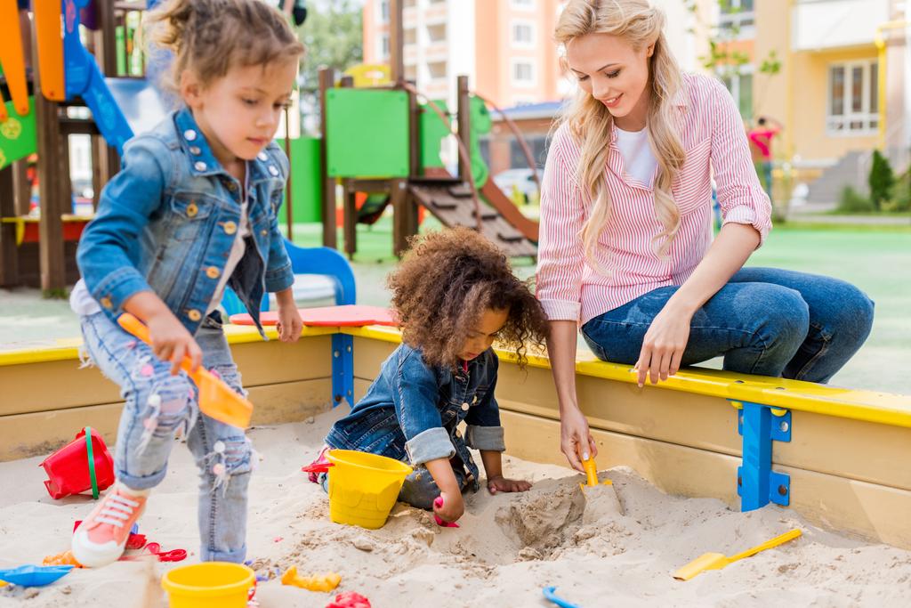 foco seletivo da mãe brincando com a filha e seu amiguinho em sandbox no playground
  - Foto, Imagem