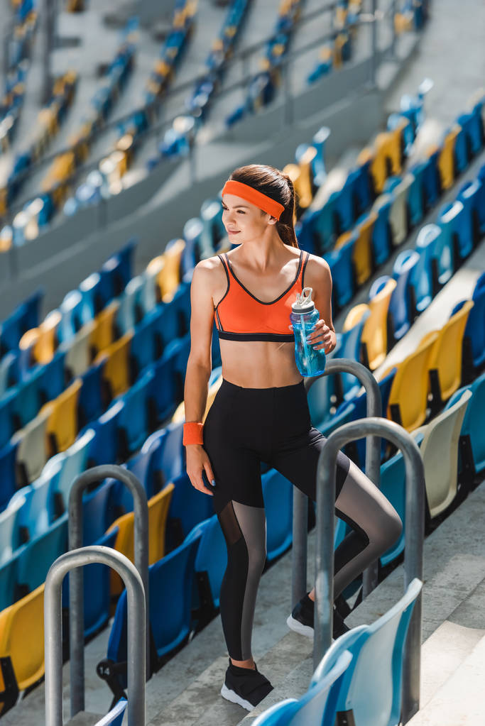 высокий угол обзора счастливой молодой женщины с бутылкой воды, стоящей на трибунах на спортивном стадионе
 - Фото, изображение