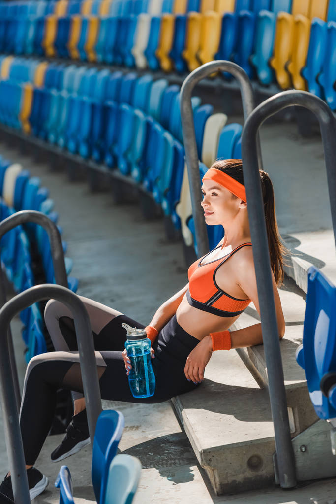 vista lateral de la mujer joven agotada con agua sentada en las escaleras en el estadio deportivo
 - Foto, imagen