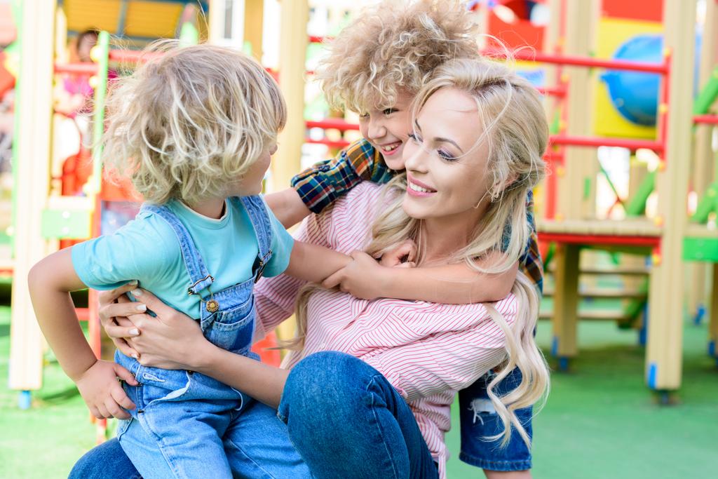 Gelukkig moeder omarmen met twee speelse schattig zonen op speelplaats  - Foto, afbeelding