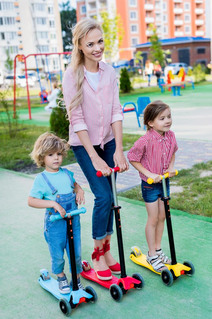 愛らしい息子と遊び場で娘と一緒にキック スクーターに乗って幸せな母  - 写真・画像