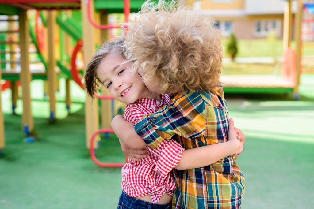 επιλεκτική εστίαση του σγουρό αγόρι φιλί και αγκαλιάζει την μικρή αδελφή στην παιδική χαρά  - Φωτογραφία, εικόνα
