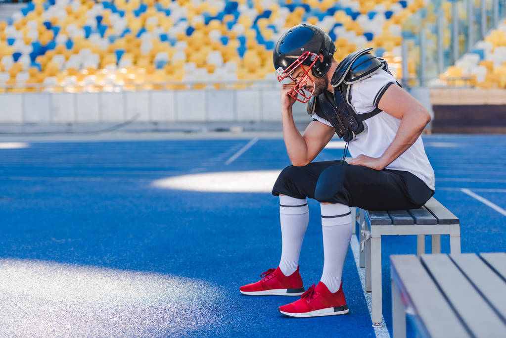 vista lateral do jovem jogador de futebol americano segurando gaiola de capacete enquanto sentado no estádio de esportes
 - Foto, Imagem