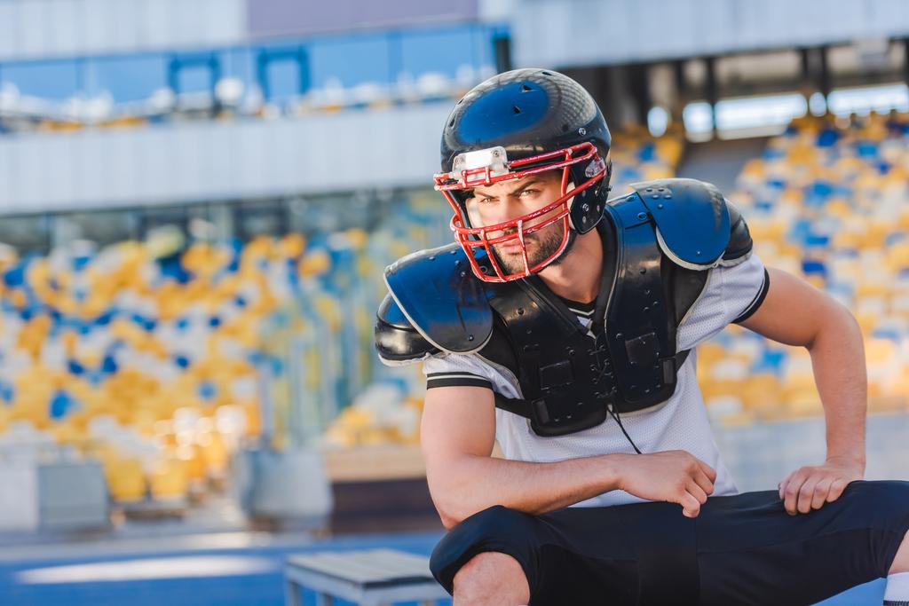 vakava nuori amerikkalainen jalkapalloilija istuu urheilustadionilla
 - Valokuva, kuva